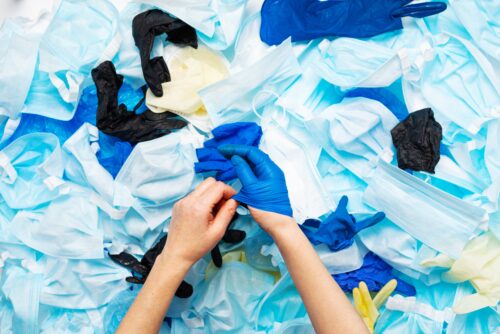 A pair of hands removes blue disposable gloves above a pile of discarded personal protective equipment.